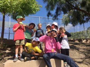 7 children and one adult in hats making goofy faces under trees in front of a chain link fence