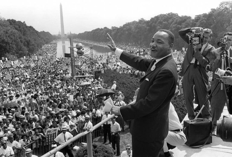 Martin Luther King Jr stands with one arm out, crowd visible in the background